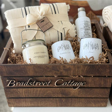  Gift basket with candles, a jar, and a towel labeled 'Bradstreet Cottage' on a blurred background.