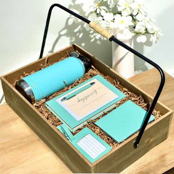 Gift set with teal 32 oz water bottle, notepad, passport journal, luggage tag, greeting card and pen in a wooden box on a light wooden surface.