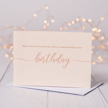  Blush pink birthday card with rose gold foil  saying Make a wish and close your eyes, dream of a happy birthday surprise. Card is on a table with fariy lights in the background