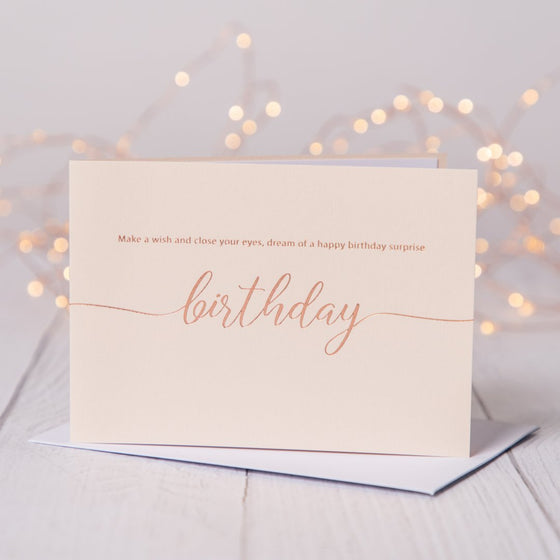 Blush pink birthday card with rose gold foil  saying Make a wish and close your eyes, dream of a happy birthday surprise. Card is on a table with fariy lights in the background